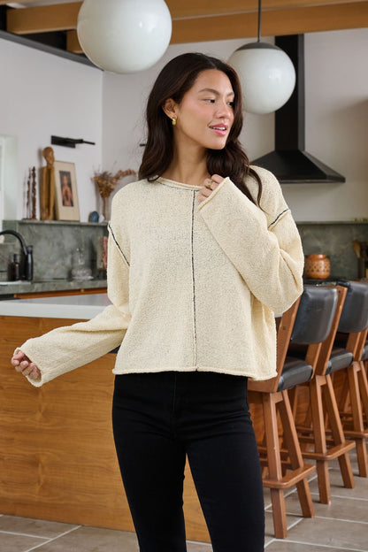 A woman wearing a Beige Contrast Stitch Detail Sweater and black jeans smiles and looks aside in a modern kitchen.