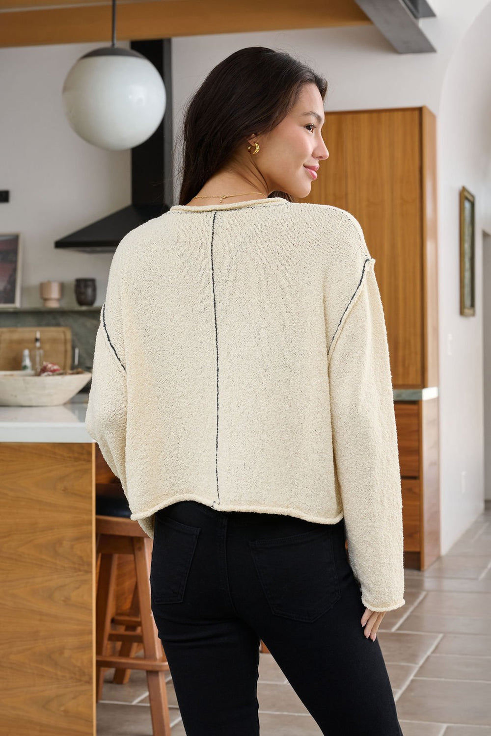 A woman with long dark hair wears the Beige Contrast Stitch Detail Sweater, standing in a modern kitchen facing away.
