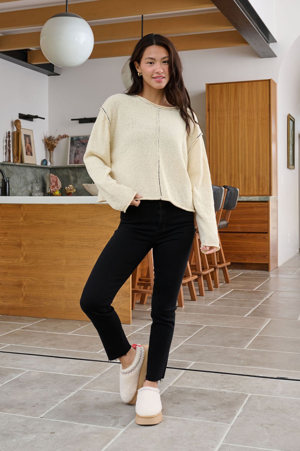 A woman wears a Beige Contrast Stitch Detail Sweater, black pants, and slippers, smiling in a modern kitchen with one hand in her pocket.