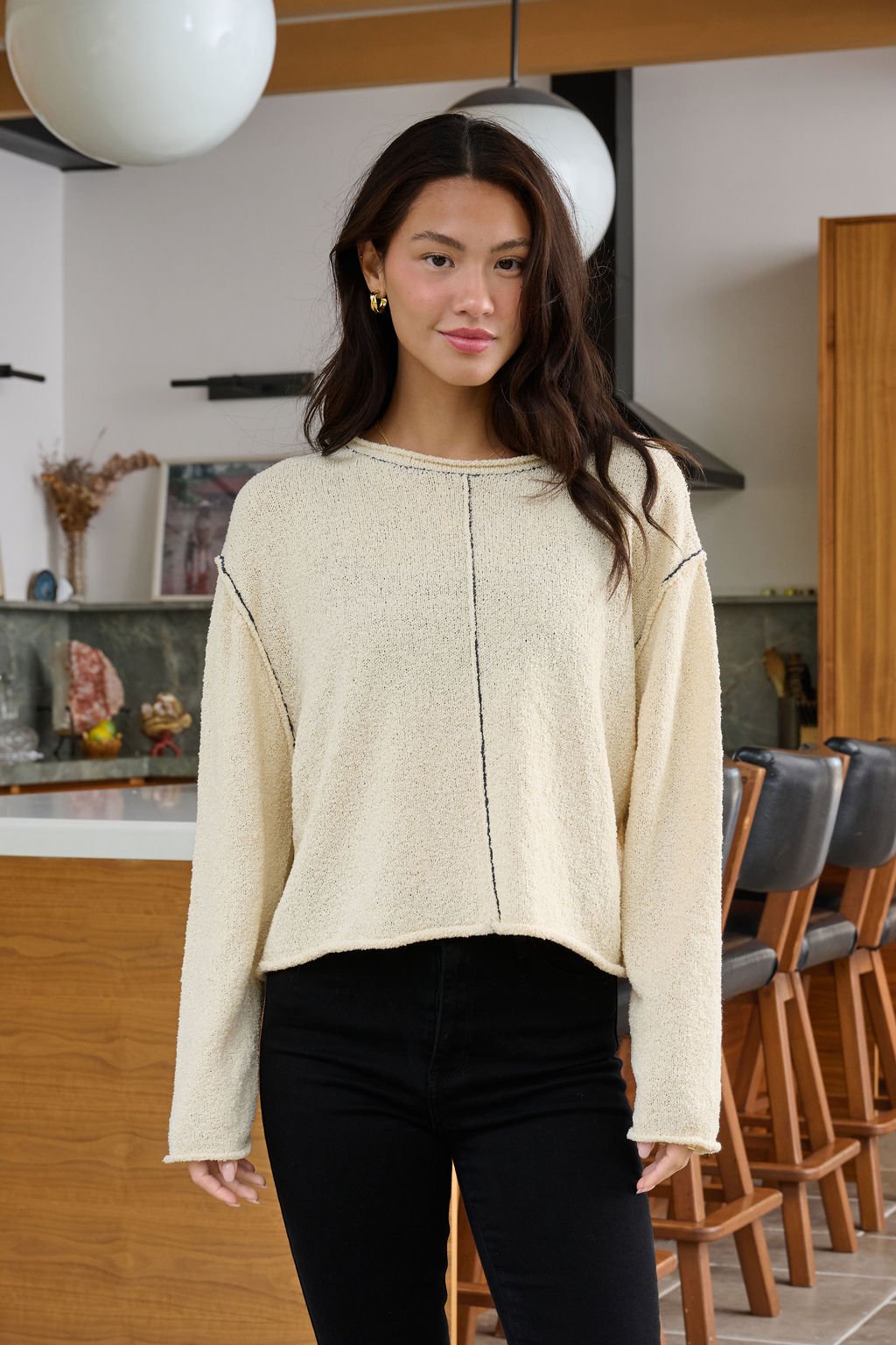A woman with long dark hair wears the Beige Contrast Stitch Detail Sweater and black pants in a modern kitchen with wooden accents.