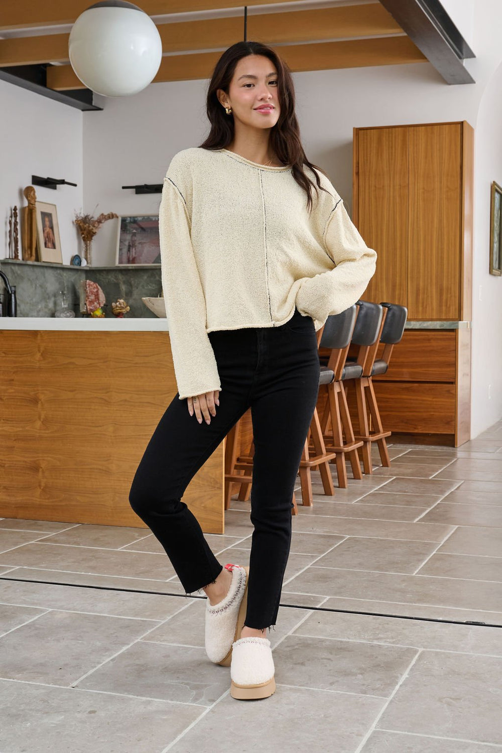 Woman stands in a modern kitchen wearing the Beige Contrast Stitch Detail Sweater - FINAL SALE, black pants, and slippers.