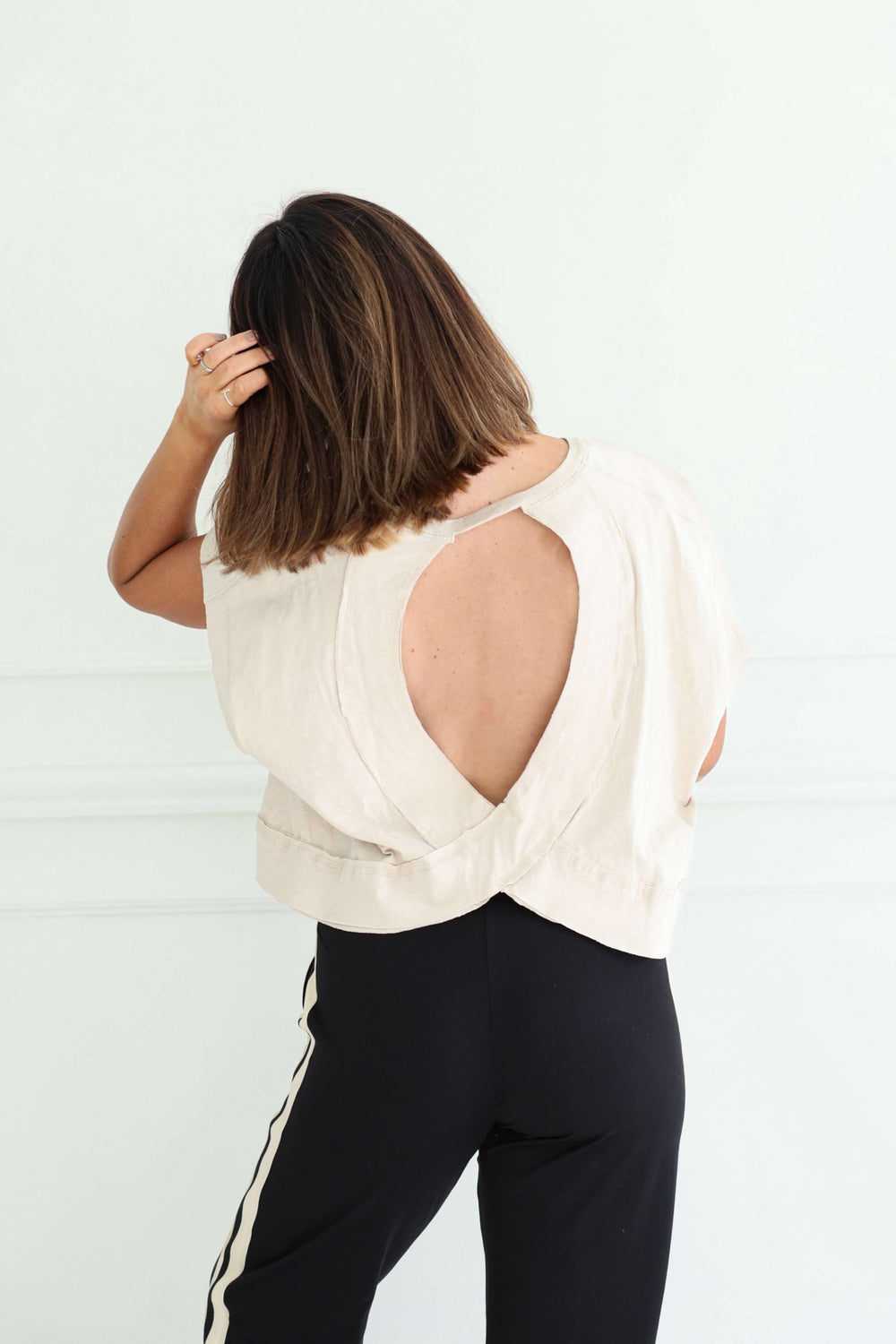 A woman with shoulder-length hair faces away, wearing the Beige Cotton Slub Open Back Top - FINAL SALE and black striped pants.