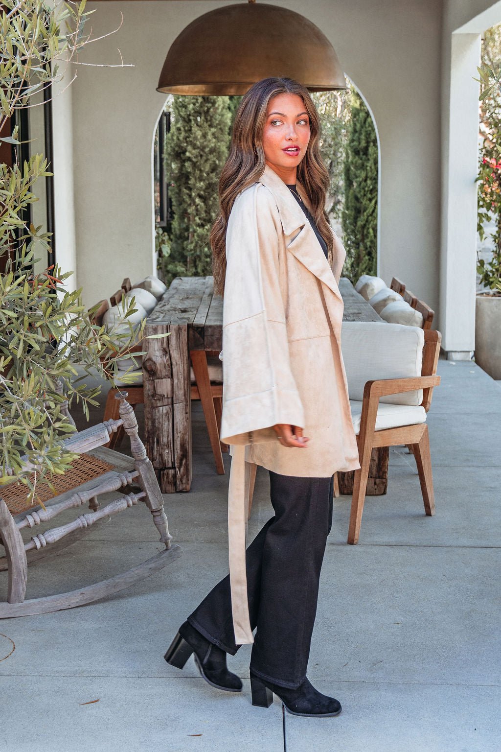 A woman wears the Beige Faux Suede Belted Coat - FINAL SALE with black pants and boots, standing near a patio dining set.
