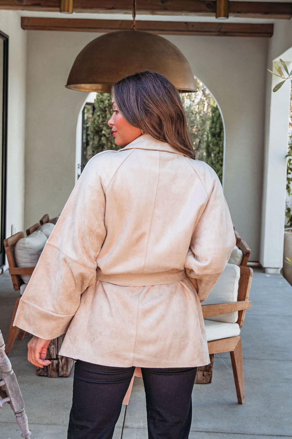 Woman with long hair outdoors in the Beige Faux Suede Belted Coat - FINAL SALE, styled with black pants near patio furniture.