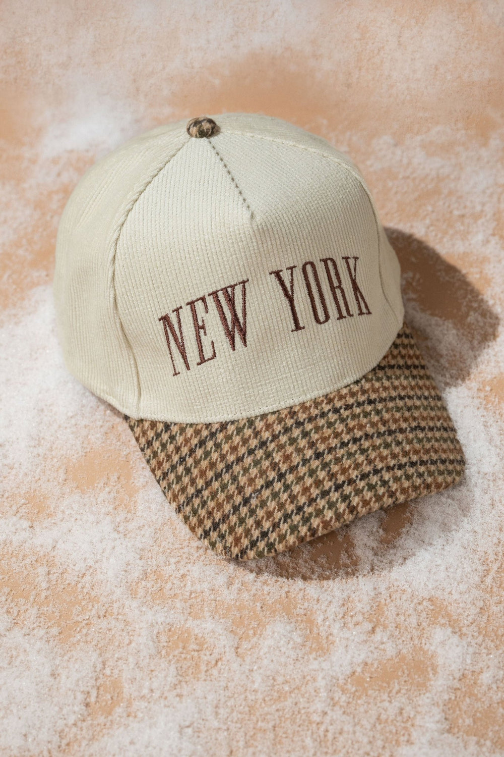 Beige New York Corduroy Baseball Hat with brown letters and houndstooth brim, styled on a sandy surface.