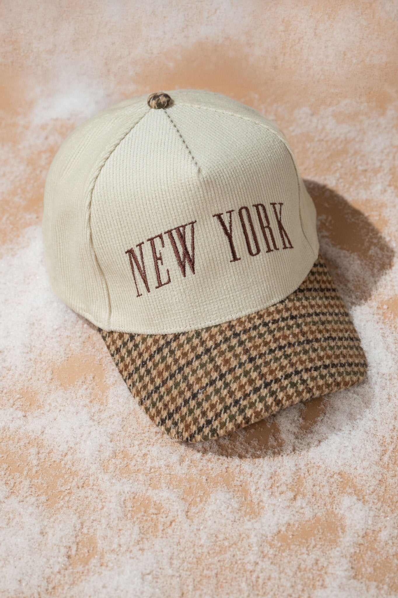 Beige New York Corduroy Baseball Hat with brown letters and houndstooth brim, styled on a sandy surface.