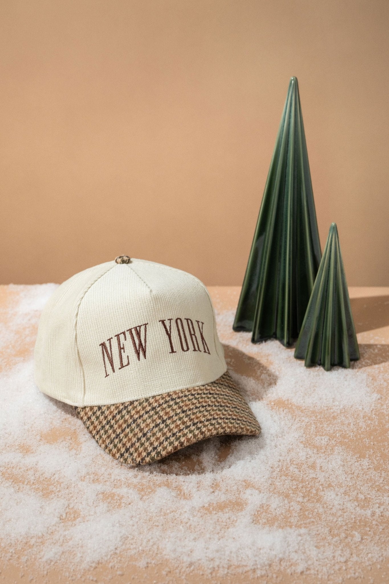 A Beige New York Corduroy Baseball Hat sits on faux snow beside green ceramic trees, set against a beige background.