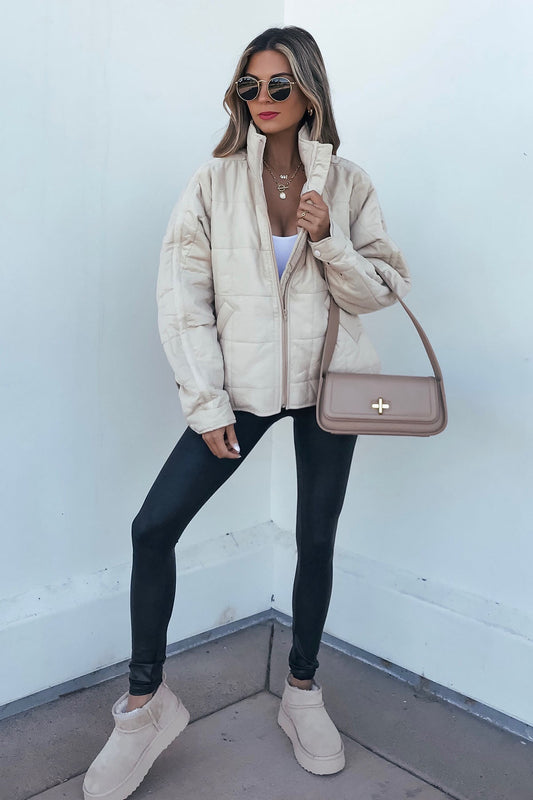 Woman in a beige quilted zip up puffer jacket, black leggings, and boots stands by a white wall—stylish winter outerwear.