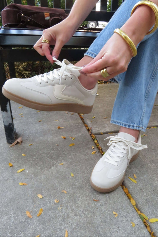 Person in blue jeans ties Beige Remus Lace Up Sneakers while sitting on a bench, yellow leaves scattered on the pavement.
