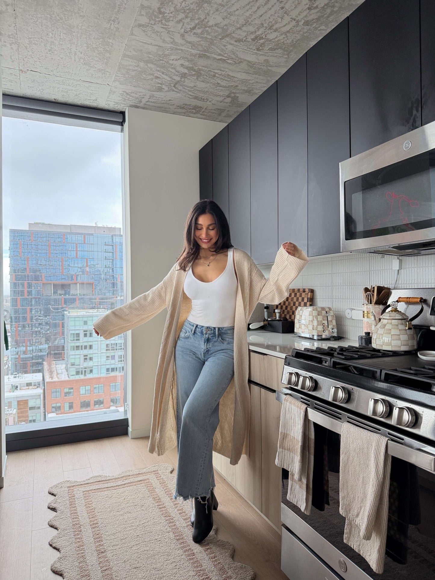 A woman smiles in a modern kitchen wearing the Beige Ribbed Open Front Duster Cardigan - FINAL SALE, ideal for winter layering.