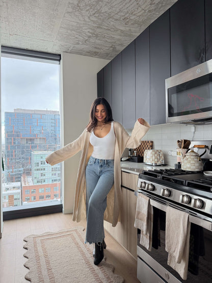 A woman smiles in a modern kitchen wearing the Beige Ribbed Open Front Duster Cardigan - FINAL SALE, ideal for winter layering.