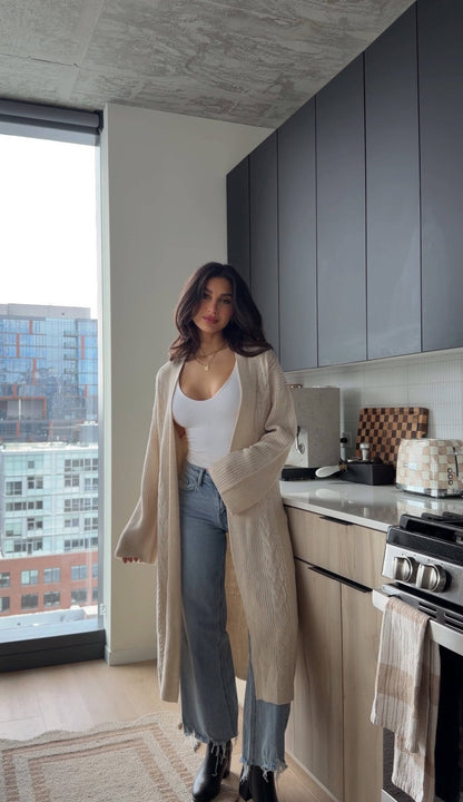 A woman wears a Beige Ribbed Open Front Duster Cardigan - FINAL SALE, a white top, and jeans in a modern kitchen with city views.