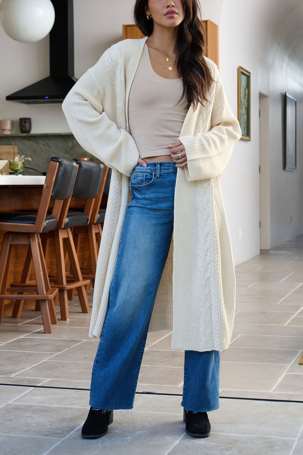 A woman in a beige ribbed open front duster cardigan, tank top, jeans, and boots stands in a modern kitchen, ideal for winter layers.