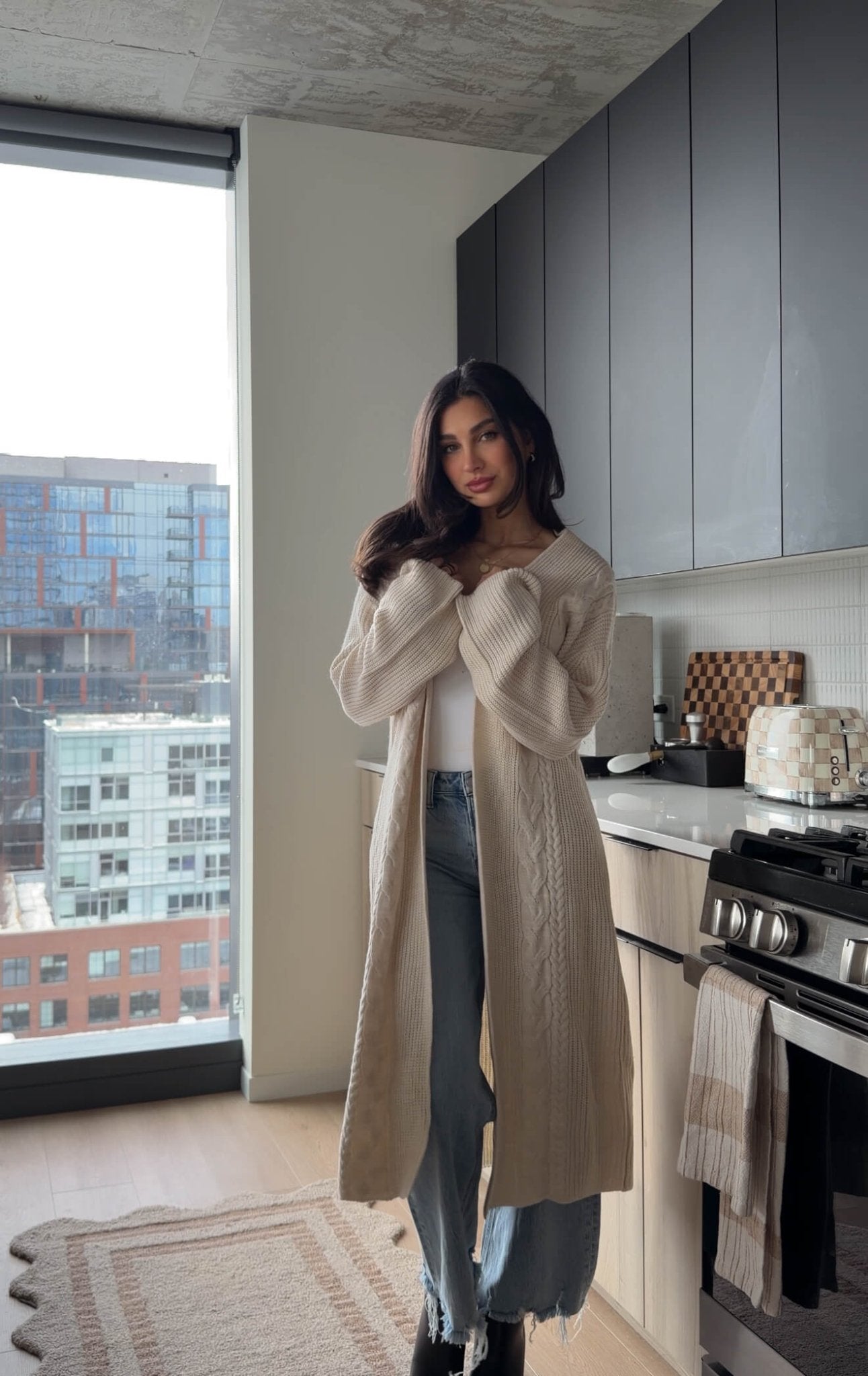 A woman wears the Beige Ribbed Open Front Duster Cardigan - FINAL SALE with jeans in a modern kitchen with city views.