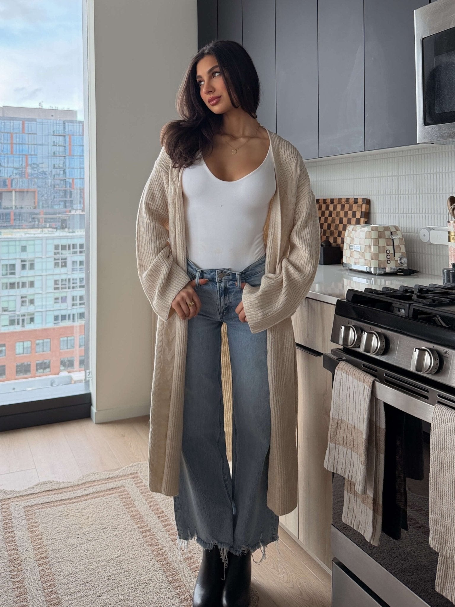 Woman wears a Beige Ribbed Open Front Duster Cardigan - FINAL SALE, white tank top, and jeans in a modern kitchen with city views.