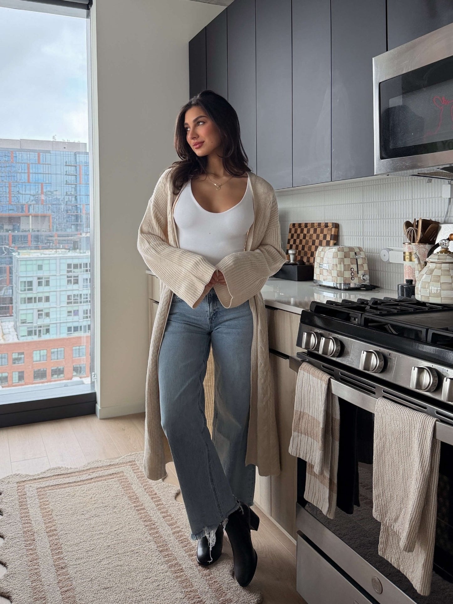 A woman in the Beige Ribbed Open Front Duster Cardigan stands by a stove in a modern kitchen, gazing thoughtfully out the window.