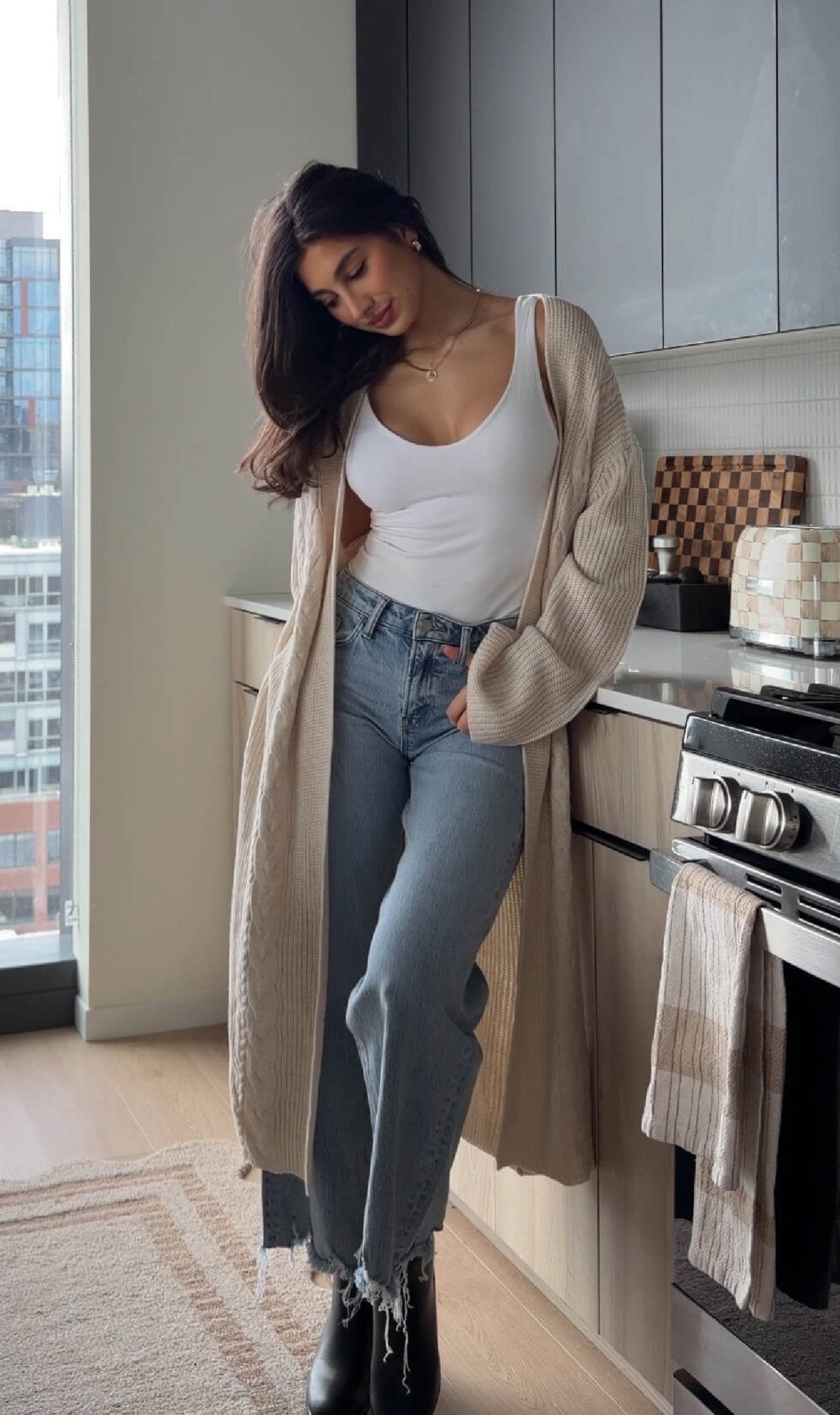 A woman wears the Beige Ribbed Open Front Duster Cardigan - FINAL SALE with a white tank and jeans in a modern kitchen by the window.