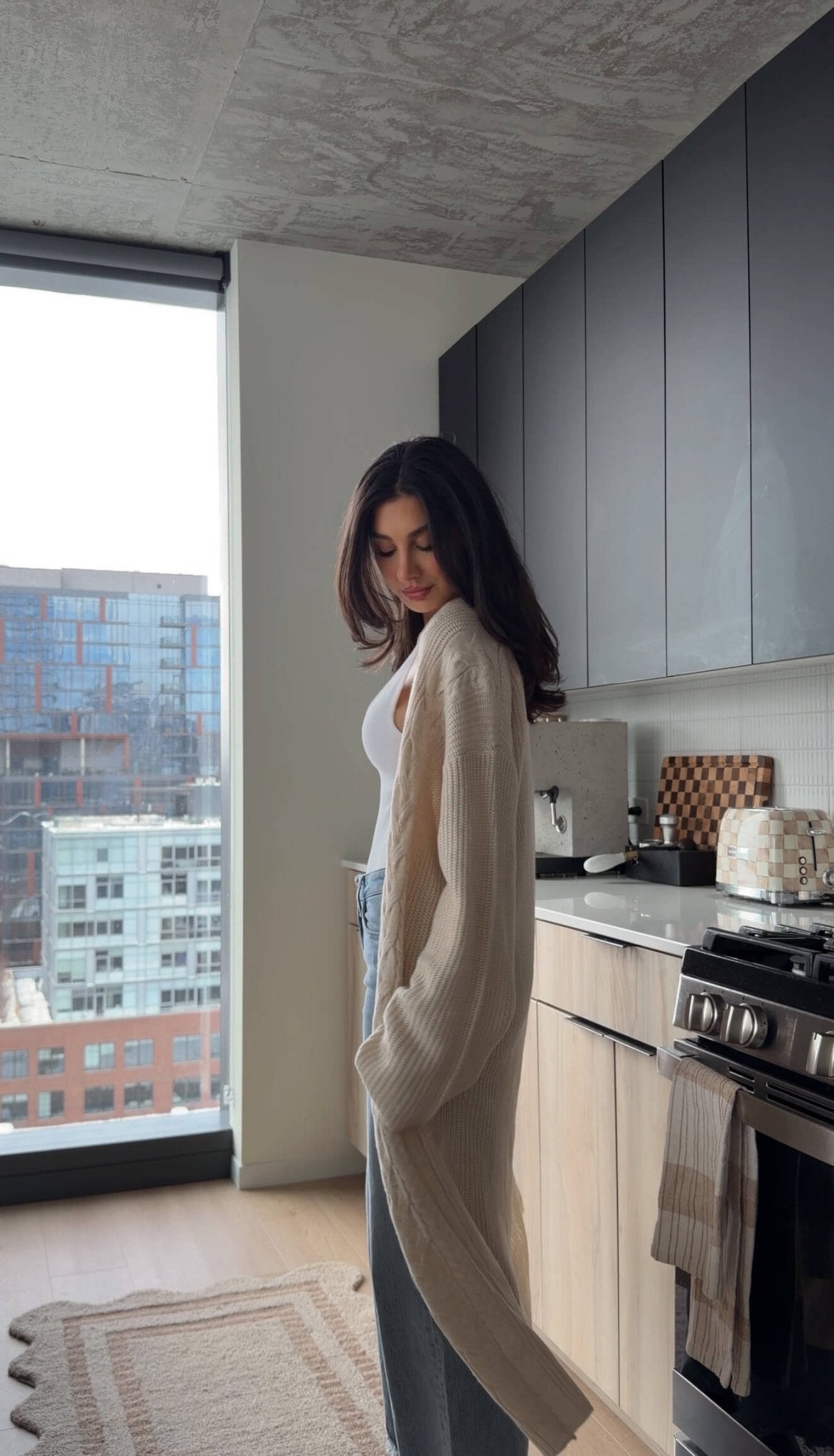 A woman wears a Beige Ribbed Open Front Duster Cardigan in a modern kitchen with city views through a large window.