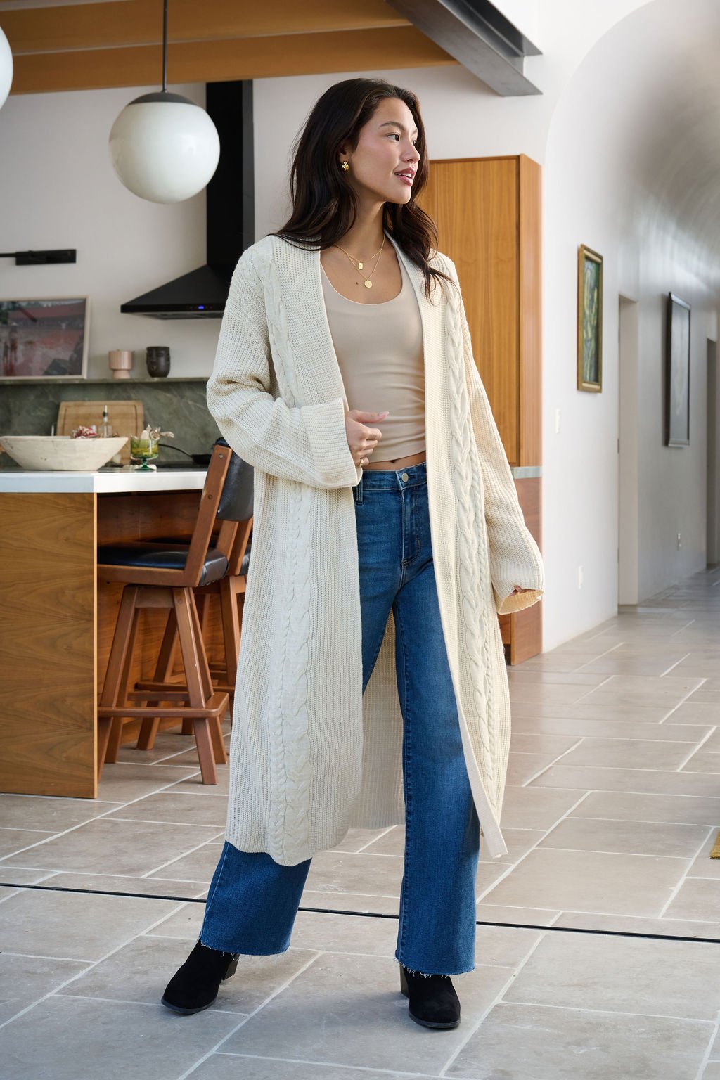 A woman wears a Beige Ribbed Open Front Duster Cardigan, tan top, and blue jeans in a modern kitchen with wood cabinets.
