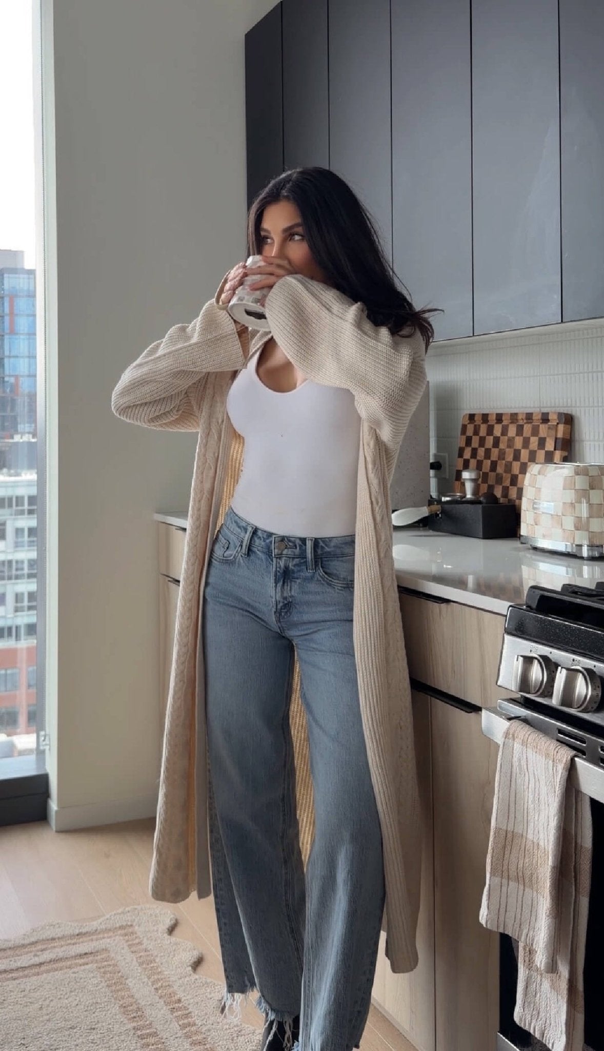 Woman in a white top, jeans, and Beige Ribbed Open Front Duster Cardigan - FINAL SALE drinks from a mug in a modern kitchen.