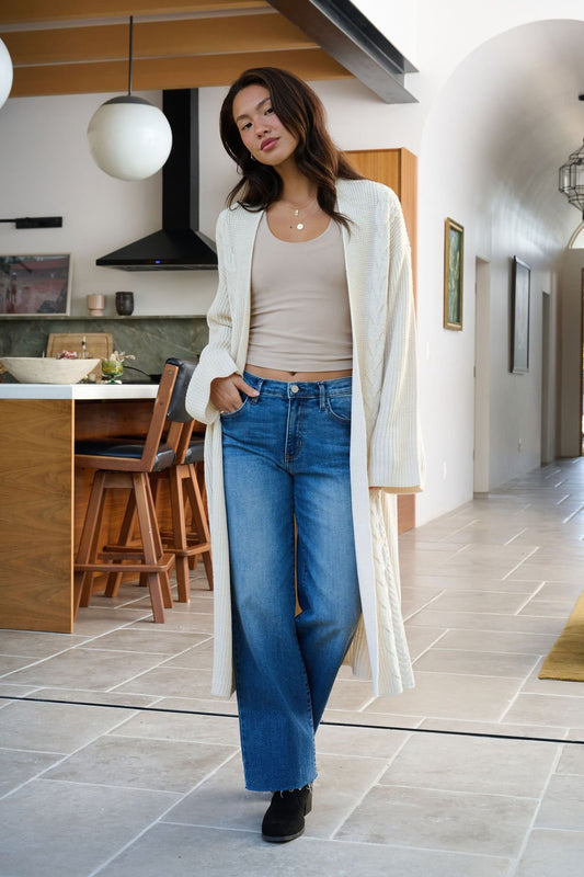 A woman wears the Beige Ribbed Open Front Duster Cardigan with blue jeans in a modern kitchen, highlighting cozy winter layering.