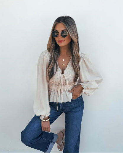 Woman in sunglasses and blue jeans poses in a Beige Ruffle Smocked Peplum Top with a v-neckline against a white wall.