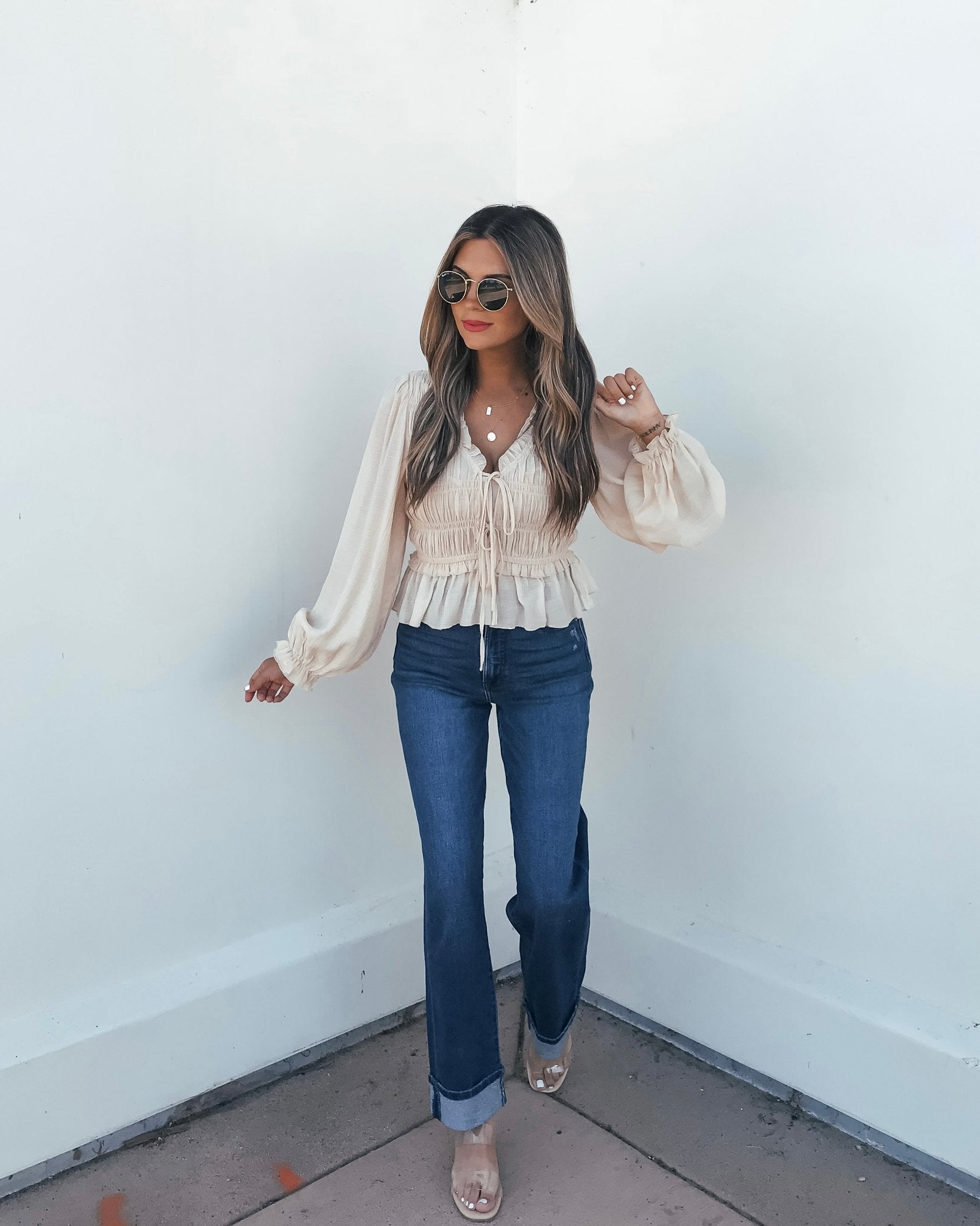 Woman in sunglasses wears a Beige Ruffle Smocked Peplum Top with v-neckline, blue jeans, and heeled sandals against a white wall.