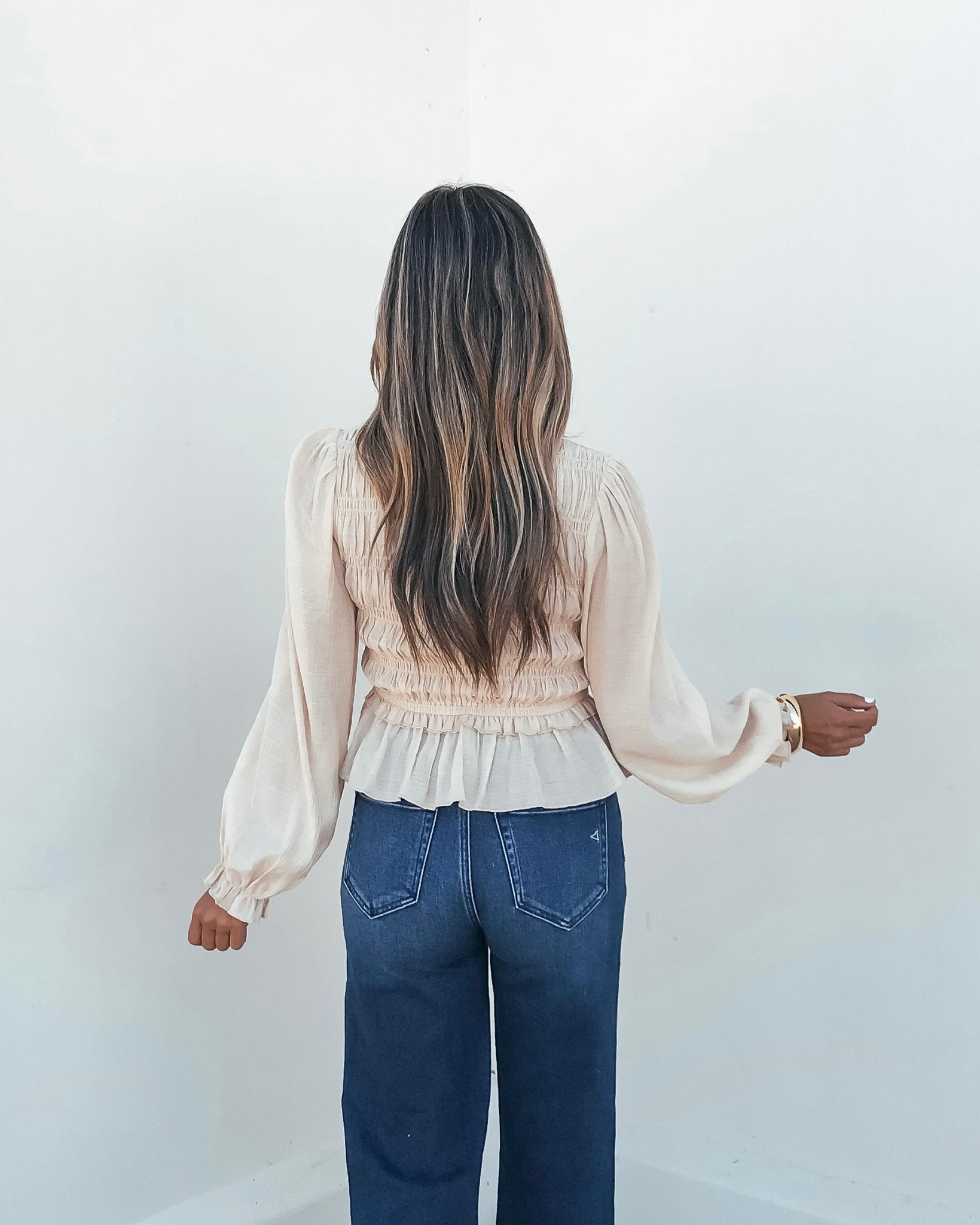 A woman with long wavy hair models the Beige Ruffle Smocked Peplum Top with blue jeans, facing a white wall.