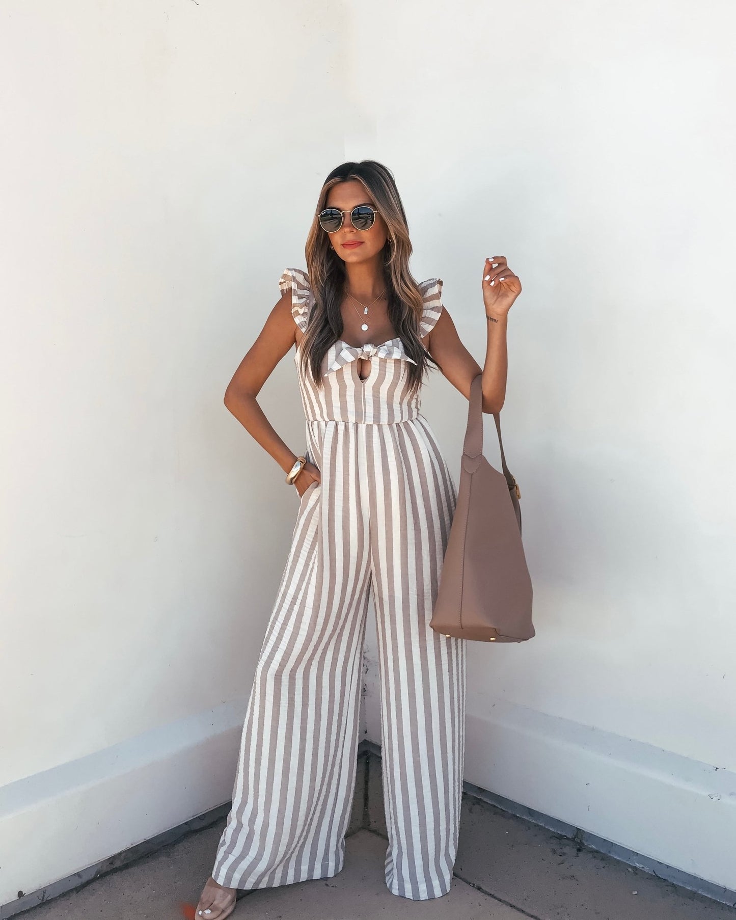 A woman models the Beige Striped Wide Leg Jumpsuit with sunglasses and a tote bag against a white wall.
