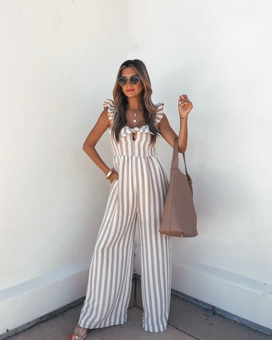 A woman models the Beige Striped Wide Leg Jumpsuit with sunglasses and a tote bag against a white wall.