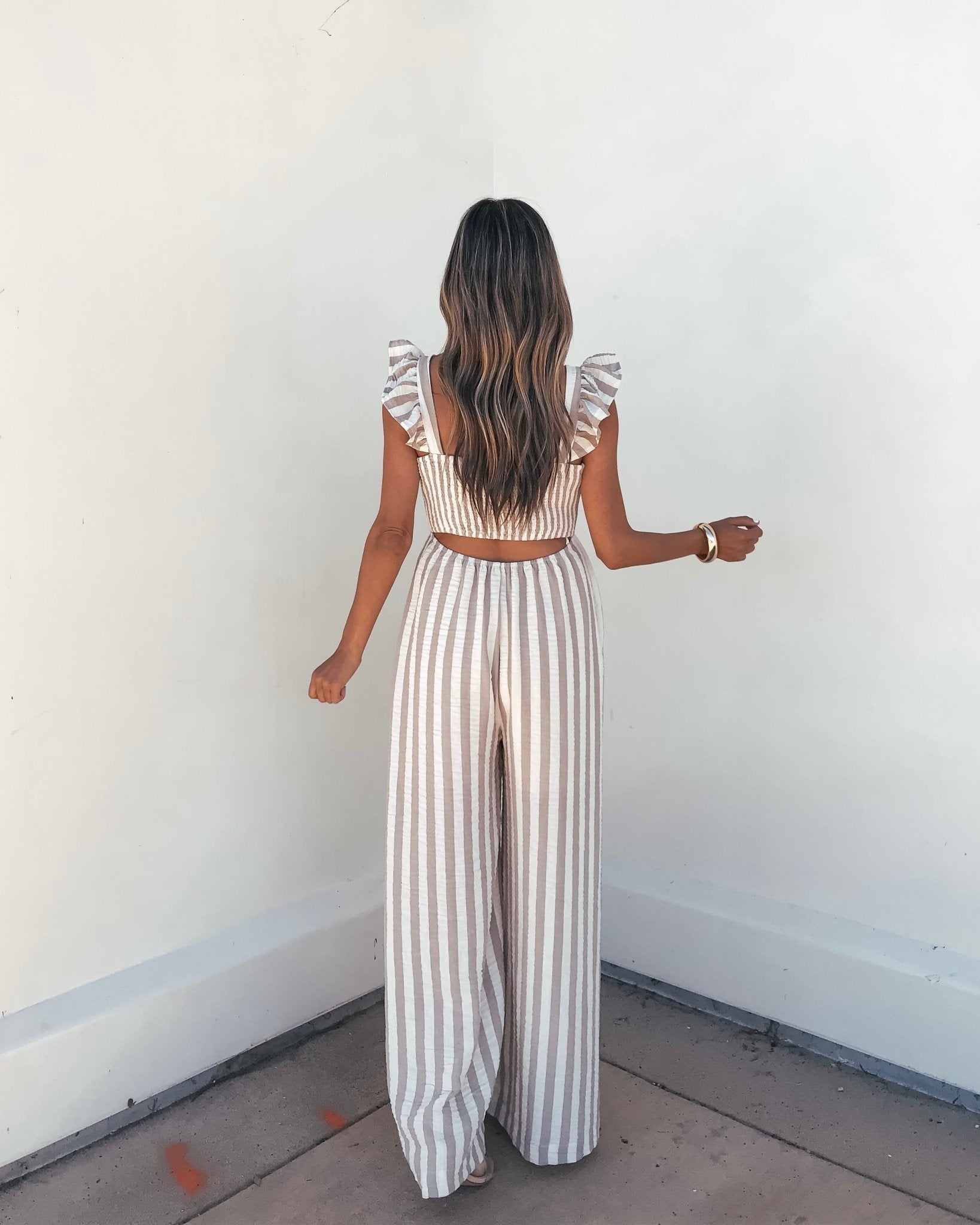 Woman with long wavy hair models the Beige Striped Wide Leg Jumpsuit, standing against a white wall, back to camera.