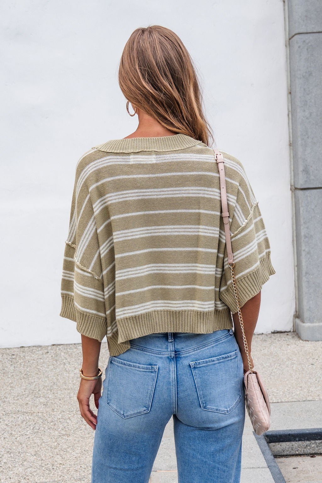 Woman with long hair in the Bellamy Striped Linen Sweater - Olive and jeans, holding a pink purse, back to camera.