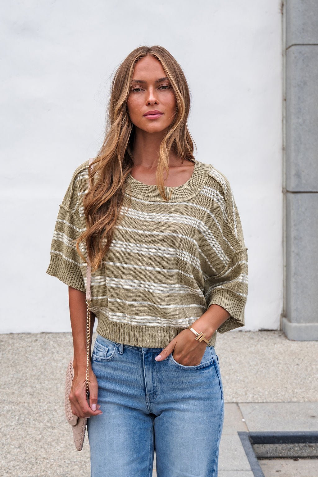 Woman with long hair wears the Bellamy Striped Linen Sweater - Olive and blue jeans, standing outdoors.