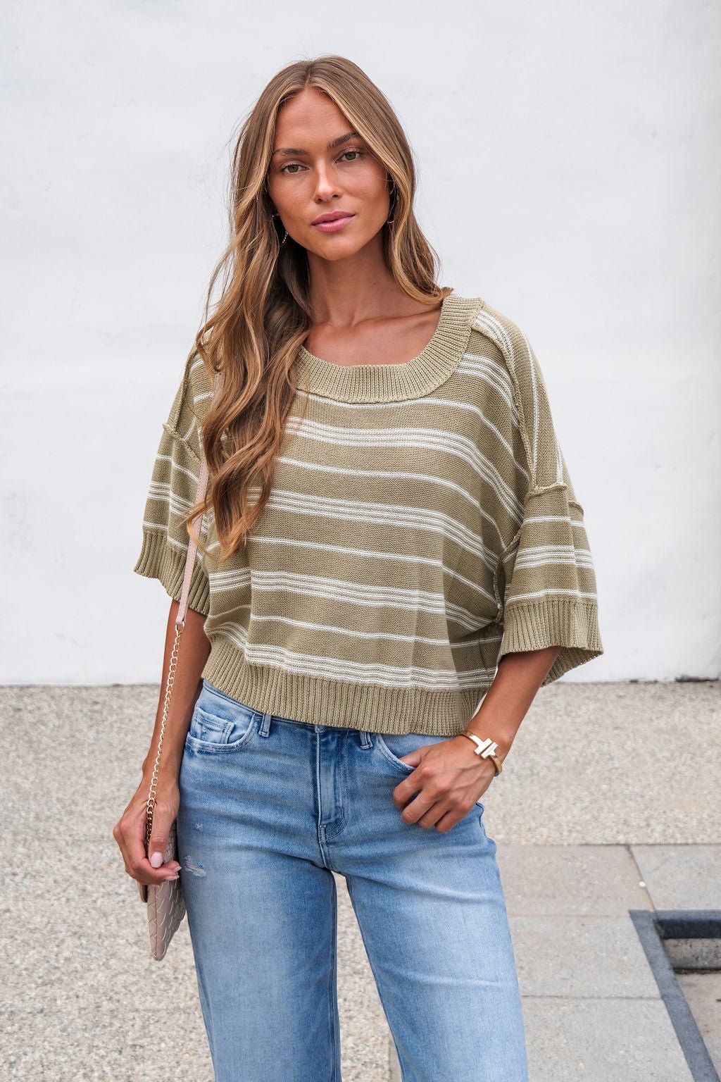 Woman in Bellamy Striped Linen Sweater - Olive and blue jeans stands outdoors holding a chain-strap purse against a white wall.