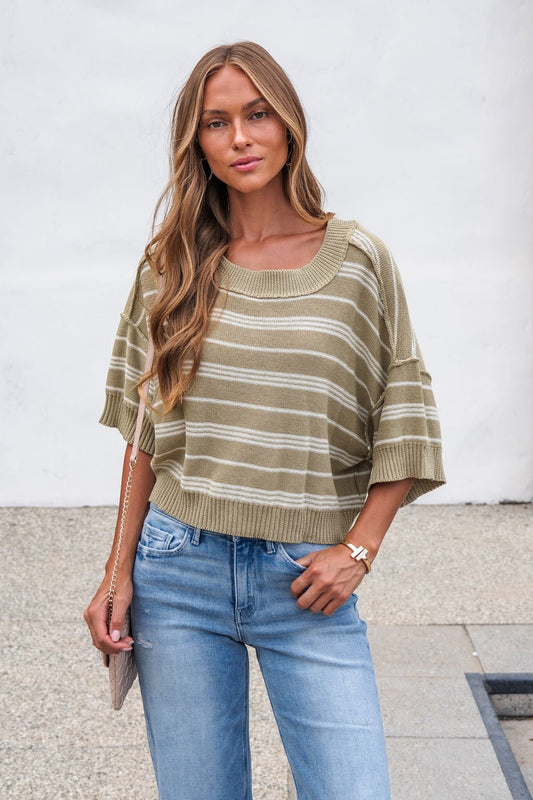 Woman in Bellamy Striped Linen Sweater - Olive and blue jeans stands outdoors holding a chain-strap purse against a white wall.