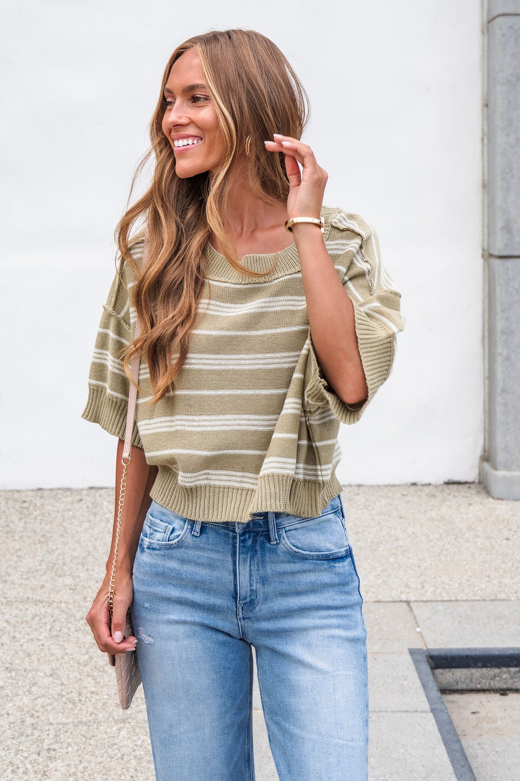 Woman smiling in the Bellamy Striped Linen Sweater - Olive and light blue jeans, holding a beige purse by a white wall outdoors.