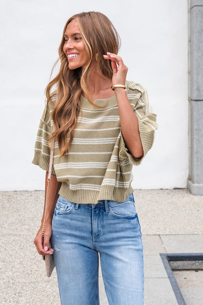 Woman smiling in the Bellamy Striped Linen Sweater - Olive and light blue jeans, holding a beige purse by a white wall outdoors.