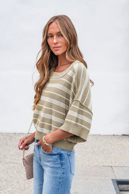 Woman with long wavy hair wears the Bellamy Striped Linen Sweater - Olive with jeans, holding a small bag outdoors.