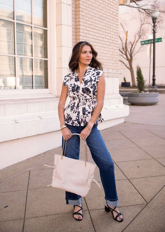 Woman in a Black and Ivory Floral Print Zip Top, blue jeans, and sandals stands on a city sidewalk with a beige purse.