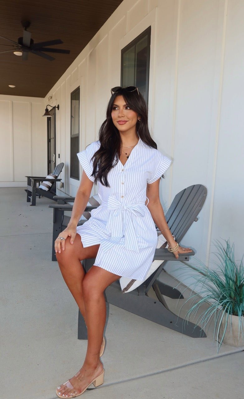 Woman in a Black and White Pinstripe Mini Dress with a collared V-neckline sits smiling on a gray chair, plant nearby.