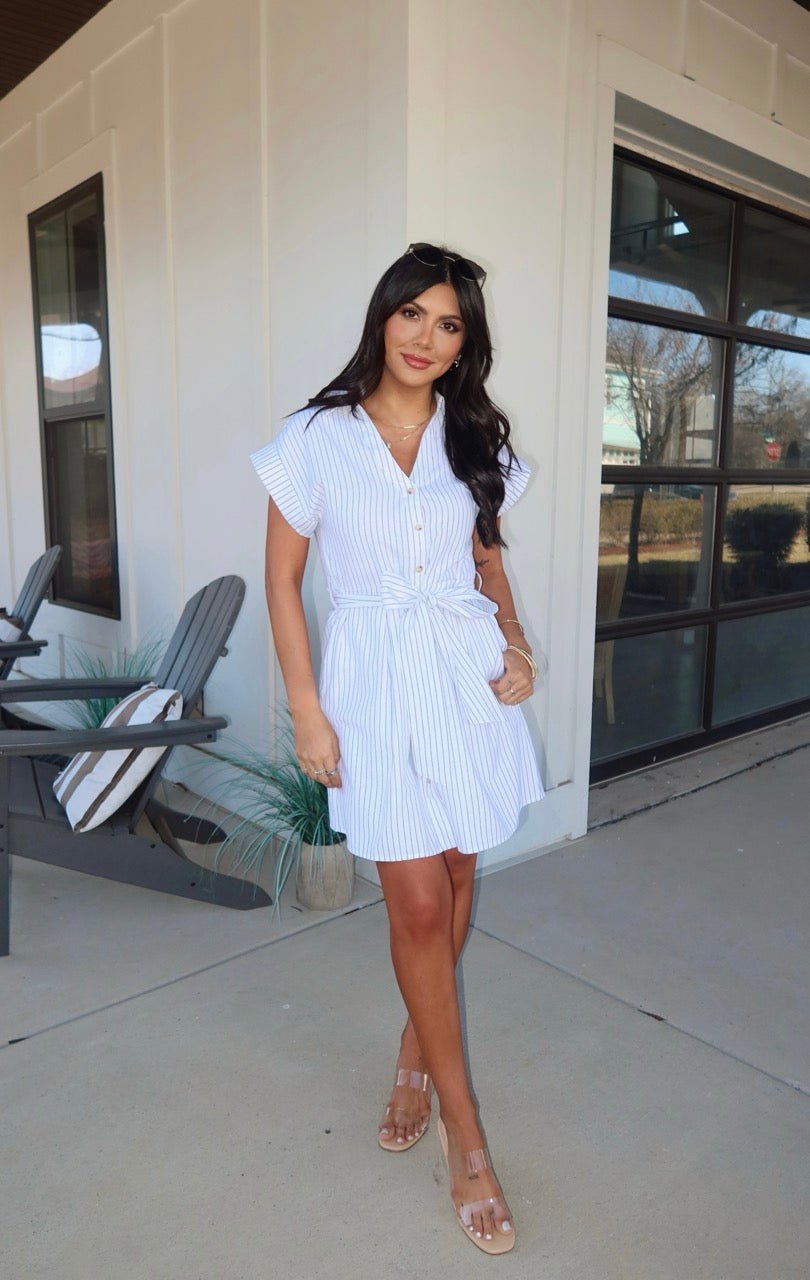 Woman standing outside a modern building in a Black and White Pinstripe Mini Dress and sandals, with long dark hair.