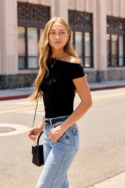Woman with long blonde hair in a Black Asymmetrical Ribbed Top and blue jeans, walking on a city street holding a small black purse.