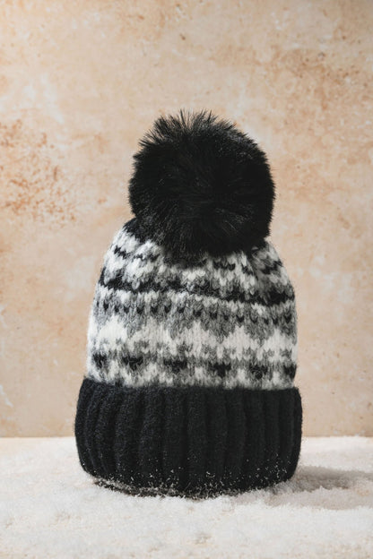 The Black Aztec Pom Beanie, featuring a black pom-pom, sits on snowy ground with a beige background.