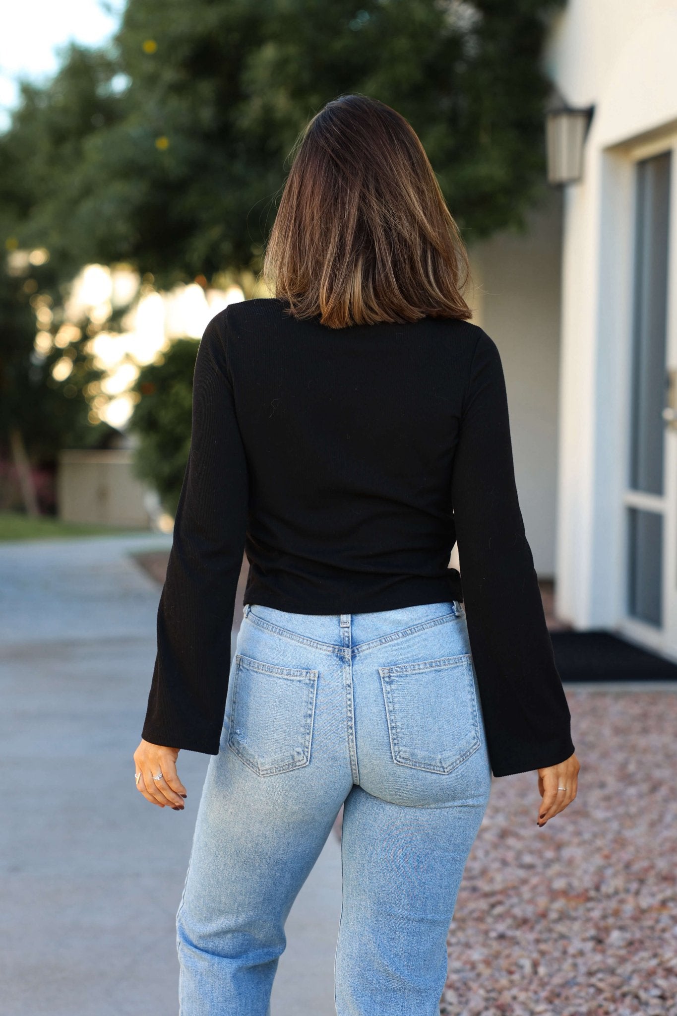 A woman wearing a Black Button Down Ribbed Top and light blue jeans stands outdoors, facing away from the camera.