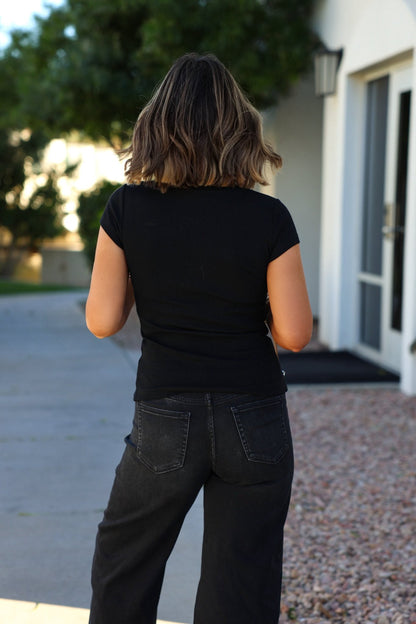 A woman in a Black Button Front Ribbed Top stands outside, facing away near a building and trees.