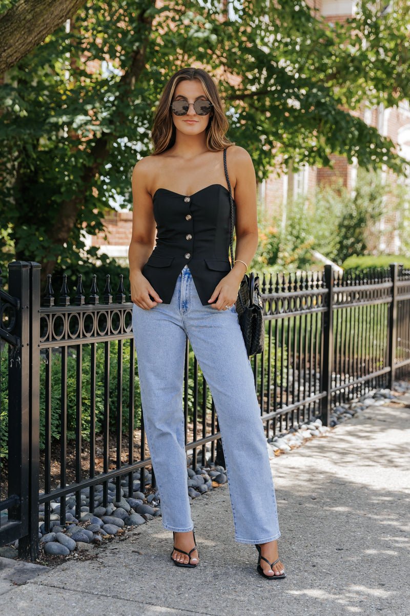 Woman outdoors in a Black Button Front Tube Top - FINAL SALE, light blue jeans, and sandals stands by a black iron fence—summer chic!.