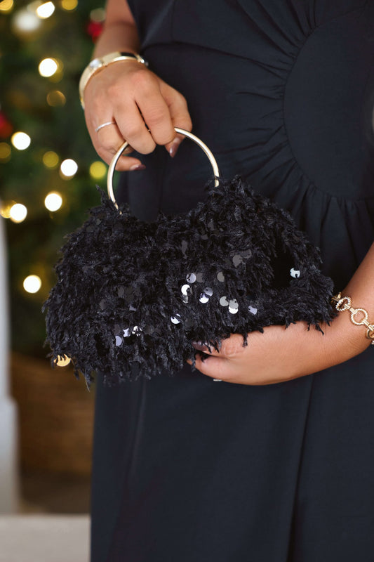 A person in a black dress holds a Black Charlotte Sequin Evening Bag with gold handles, framed by soft holiday lights.