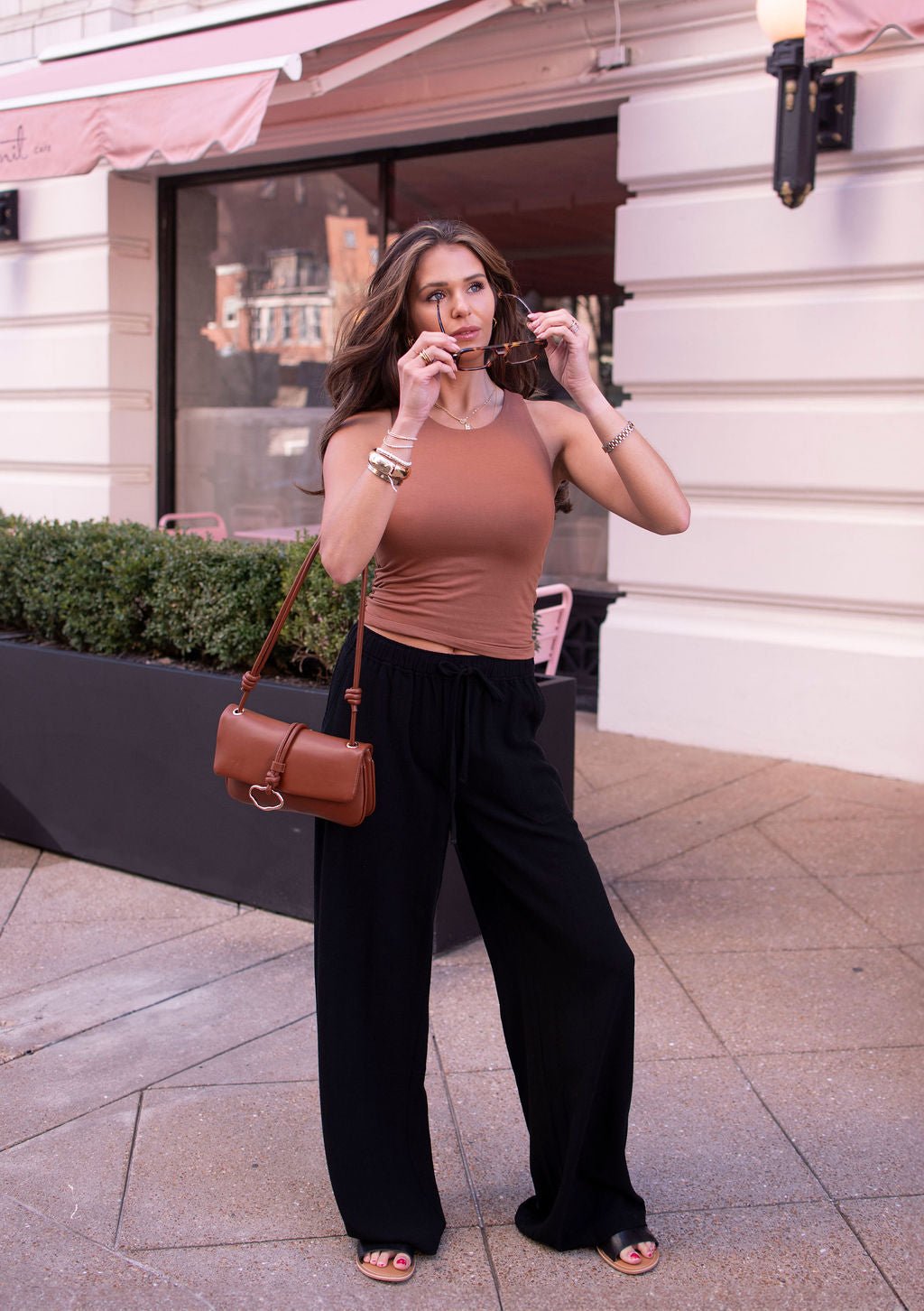 Woman in black drawstring wide leg linen pants and a brown tank top stands outside with sunglasses and a brown purse.