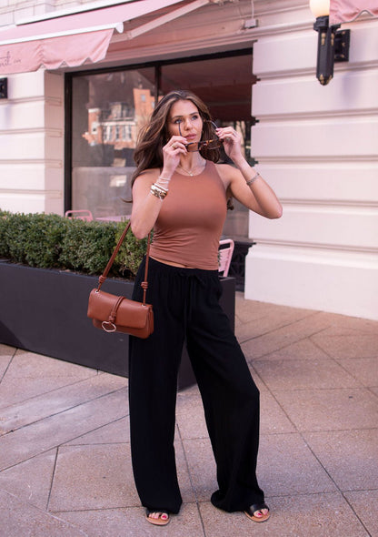 Woman in black drawstring wide leg linen pants and a brown tank top stands outside with sunglasses and a brown purse.