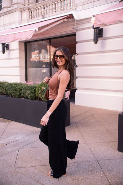 Woman with long brown hair wears sunglasses, a brown tank top, and Black Drawstring Wide Leg Linen Pants, smiling outdoors by a white building.